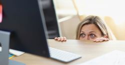 Woman hiding behind her desk and peeking at her computer screen in fear